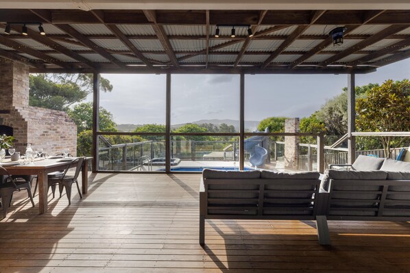 A covered outdoor area with a wooden deck, a brick fireplace, a dining table with chairs, a pool, and a seating area with cushions.