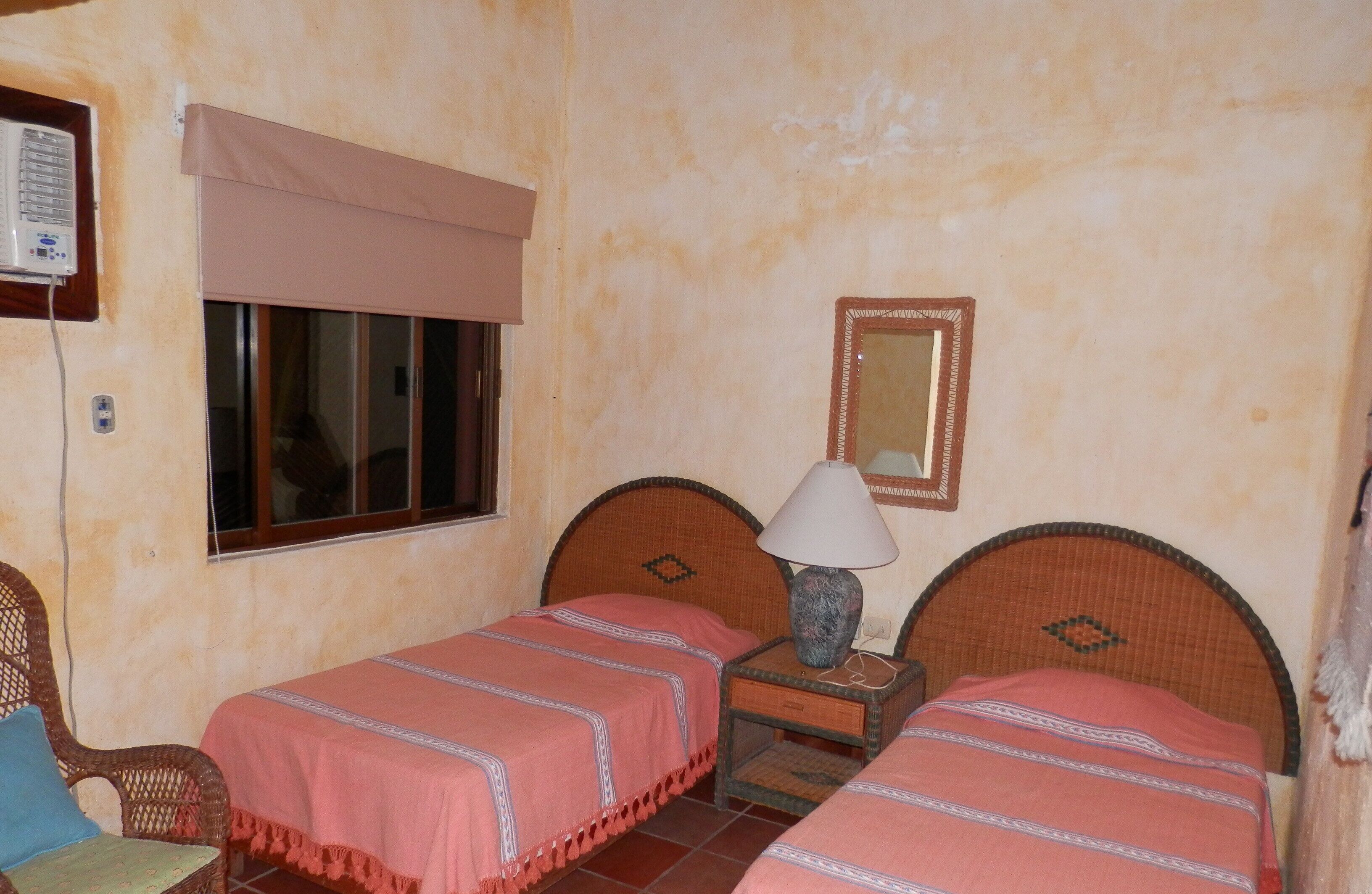 Een kamer met twee enkele bedden, een nachtkastje met lamp, een raam met jaloezieën en een luchtverkoeler aan de muur.