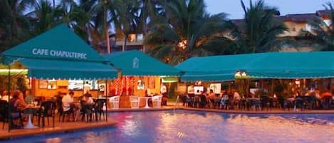 A poolside restaurant and bar with outdoor seating, palm trees, and a pool.