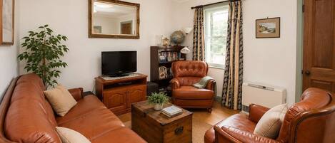 Uma sala de estar com um sofá de couro marrom, uma poltrona de couro marrom, uma mesa de centro de madeira, uma televisão sobre um móvel de madeira, uma estante de livros, um vaso de plantas e uma janela com cortinas.