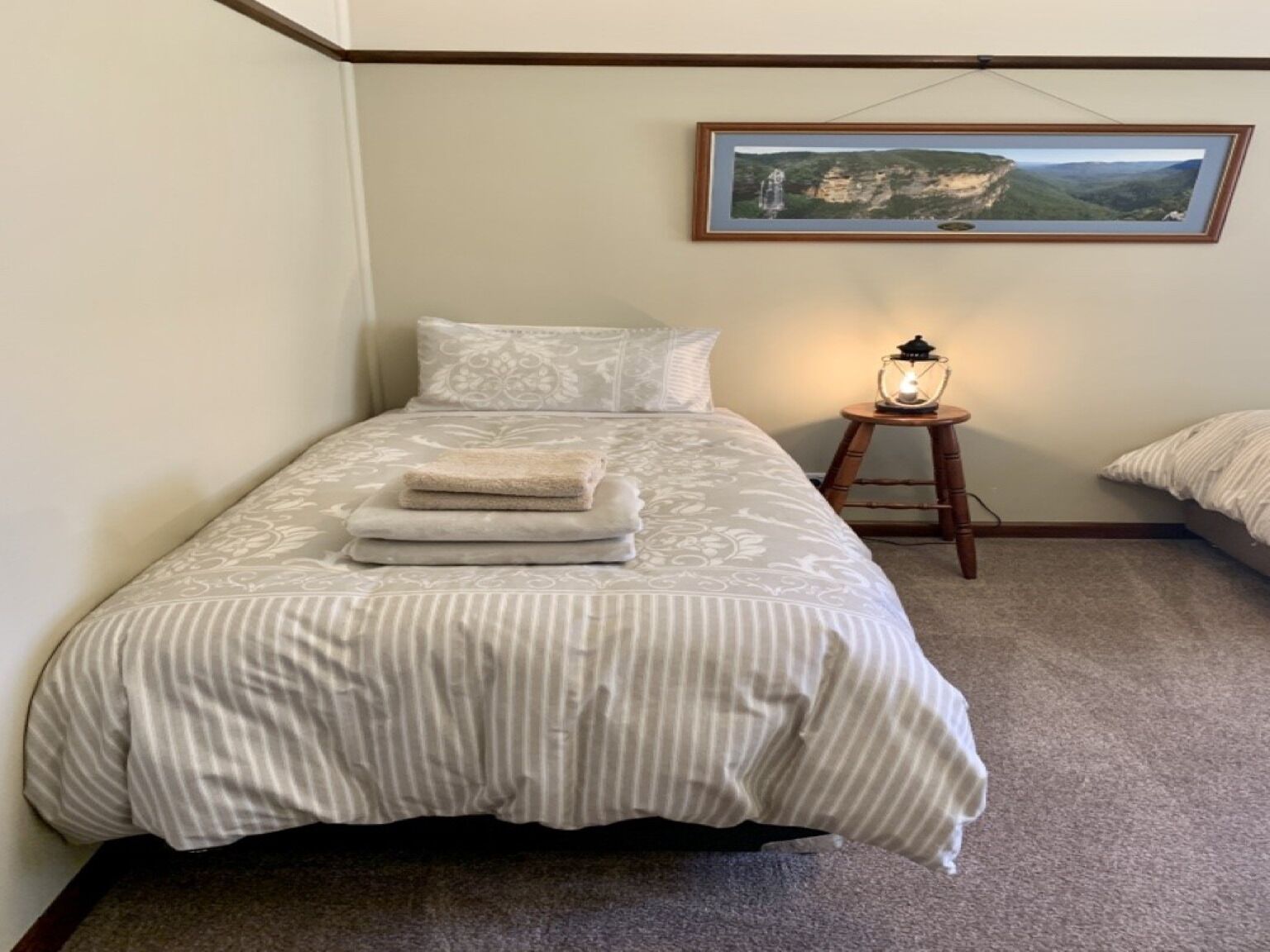 Ein ordentlich bezogenes Bett mit gestreifter Bettdecke und Kissen, ein kleiner Nachttisch mit einer Laterne und ein gerahmtes Landschaftsbild an der Wand.