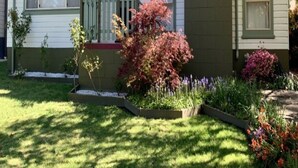 Una casa de un piso con un porche delantero, un pequeño jardín con diversas plantas y un cielo azul despejado.