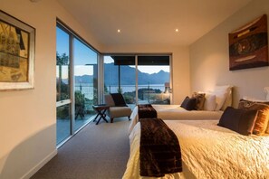 A modern bedroom with a large bed, a sofa, and a chair. It features a balcony with a view of mountains and a body of water.