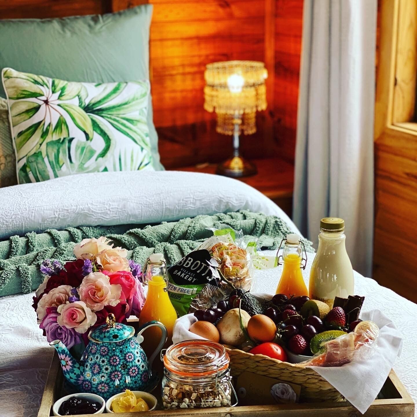 Un plateau de petit-déjeuner joliment préparé, avec des fleurs, du pain, des œufs et une bouteille de jus, posé sur un lit.