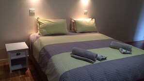 Een net opgemaakt bed met een groen-paars geruit laken, twee kussens en een witte nachtkastje met een lade.
