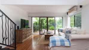 Een ruime woonkamer met een houten vloer, een zithoek, een televisie en uitzicht op een weelderige tuin door grote glazen schuifdeuren.