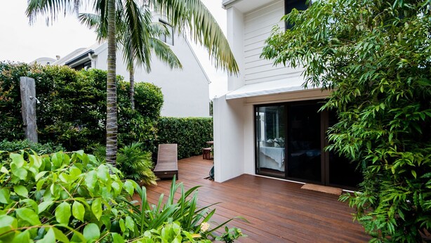 Een modern huis met een houten terras, een overdekte zithoek en weelderige begroeiing.