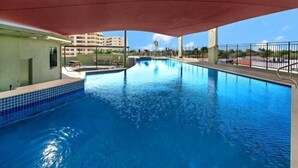 An indoor swimming pool with clear blue water, a covered area, and a view of a building in the background. - Nomiki across the avenue (parap)