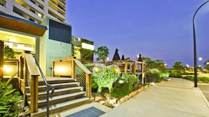 A hotel entrance with a staircase, a covered area with plants, and a well-lit pathway. - Nomiki across the avenue (parap)