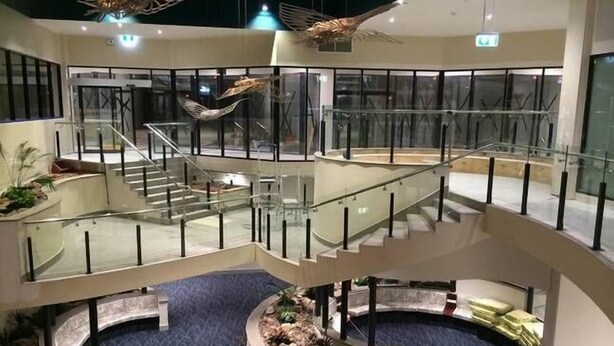A multi-level interior with glass railings, a central atrium with a circular seating area, and decorative metal sculptures. - Nomiki across the avenue (parap)