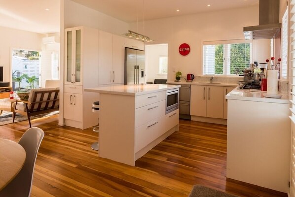 A modern kitchen with white cabinetry, a central island, and a dining area with a wooden floor. - Oneroa Village, 5 mins to Oneroa beach, Restaurants & Shops | Be My Guest (Oneroa)