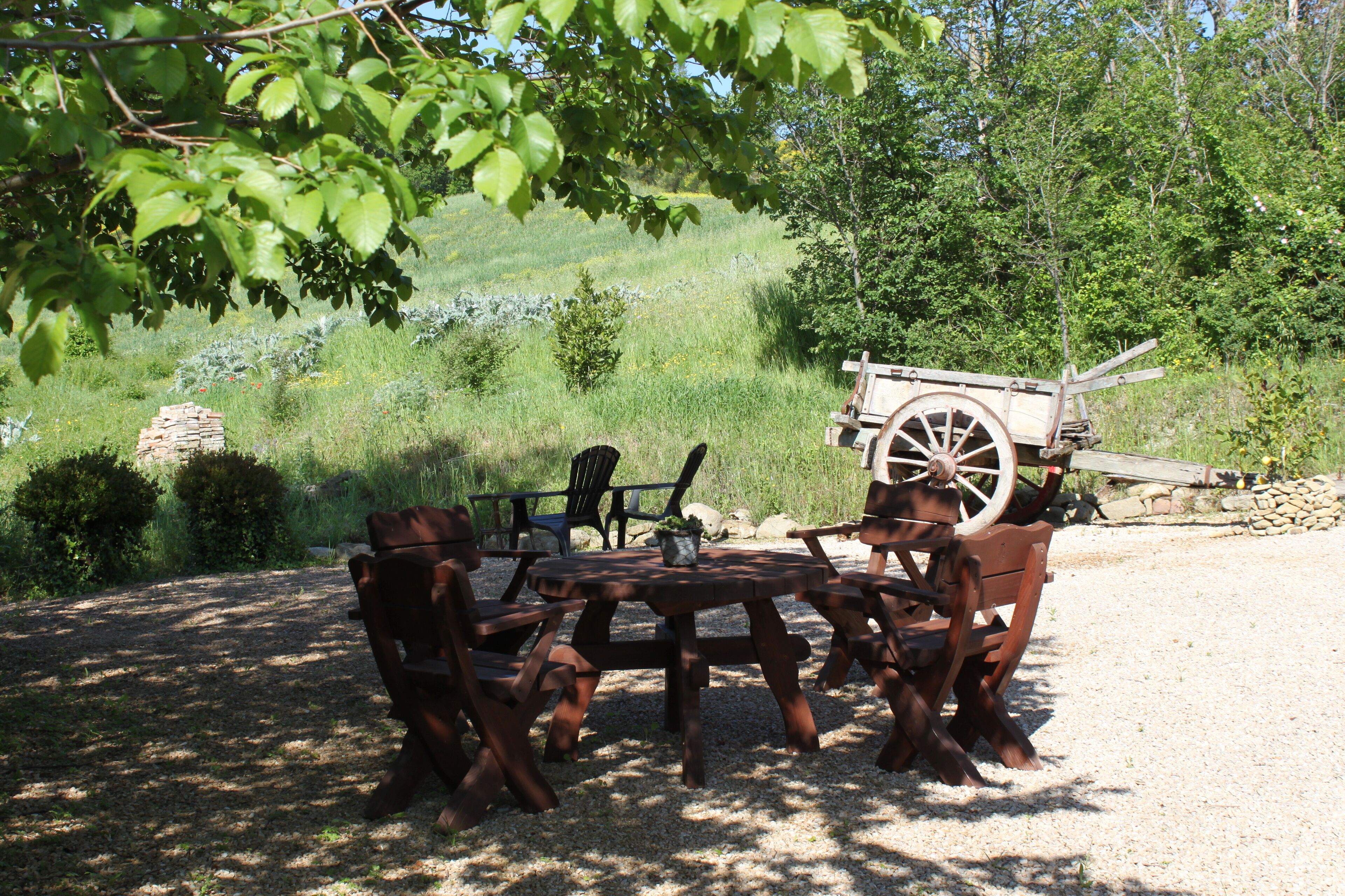 A rustic outdoor seating area with a wooden table and benches, surrounded by greenery and a stone wall.