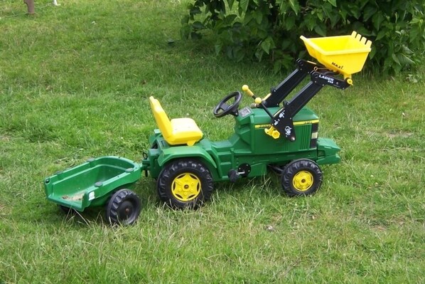 A green toy tractor with a yellow bucket and a green trailer on a grassy field.
