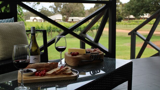 Une table avec une bouteille de vin, un verre de vin, un bol de cerises, une planche à découper avec du fromage et de la charcuterie, et un panier de pain.