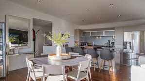Una cocina moderna con electrodomésticos de acero inoxidable, un comedor con una mesa redonda y sillas blancas, y un espejo grande sobre la mesa.