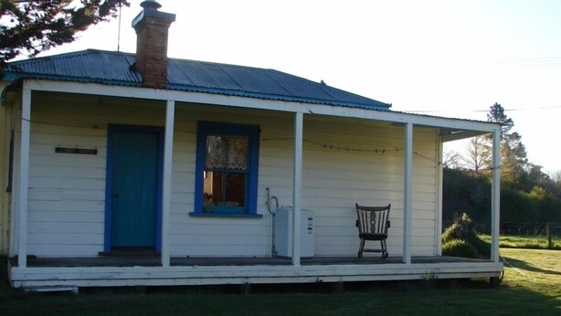 A single-story house with a blue door and window, a chimney, and a porch with a bench. - Quaint character cottage on working farmlet, Running Sheep & Beef. (Upper Moutere)