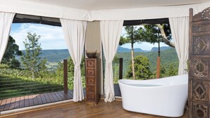 A room with a large bathtub, wooden cabinet, and curtains overlooking a scenic landscape. - Clouds Safari - Glamping with views (Beechmont)