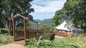 A wooden gate with a decorative headpiece, a wooden fence, and a tent-like structure in a lush, green landscape with mountains in the background. - Clouds Safari - Glamping with views (Beechmont)