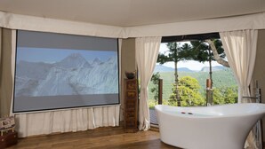 A room with a large screen displaying a mountain view, a white bathtub, and a wooden cabinet. - Clouds Safari - Glamping with views (Beechmont)