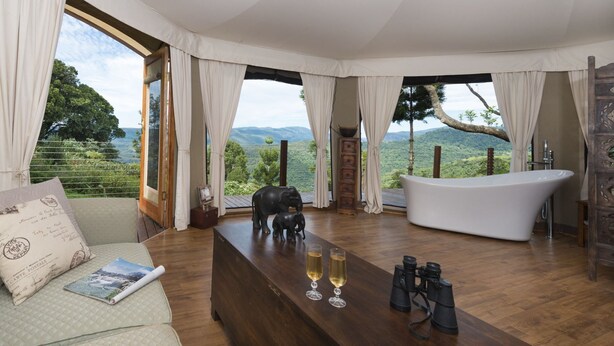 A room with a large bathtub, a wooden table with two glasses and binoculars, a sofa with a magazine, and a view of a mountainous landscape through large windows. - Clouds Safari - Glamping with views (Beechmont)