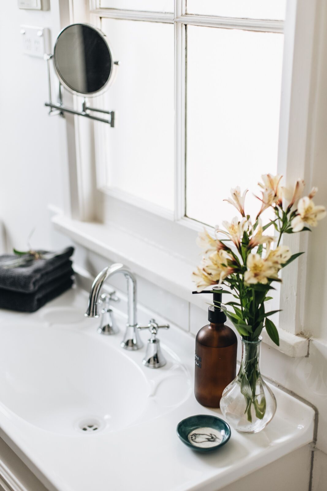 Ein Badezimmer mit einem weißen Waschbecken, einem Spiegel, einem Fenster und einer Vase mit Blumen.