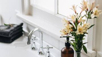 Ein Badezimmer mit einem weißen Waschbecken, einem Spiegel, einem Fenster und einer Vase mit Blumen.
