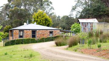 A stone house with a metal roof, surrounded by a fenced yard and a dirt road.