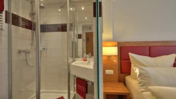 Una habitación de hotel moderna con ducha envuelta en vidrio, mesita de noche de madera, cama con cabecera roja y lavamanos blanco.