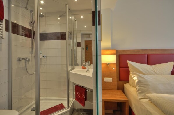 Una habitación de hotel moderna con ducha envuelta en vidrio, mesita de noche de madera, cama con cabecera roja y lavamanos blanco.