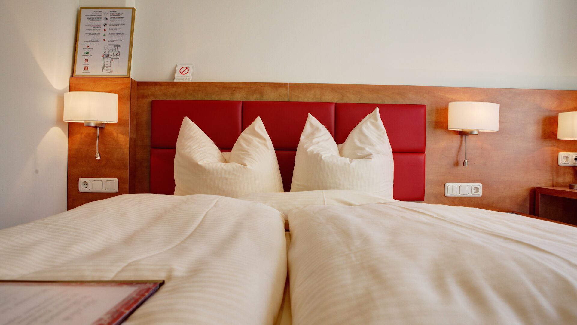 A hotel room with a large bed, two bedside lamps, a red headboard, and a wall-mounted information board.