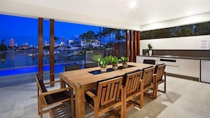 A modern dining area with a wooden table and chairs, a view of a cityscape and water body, and a kitchen area with white cabinets and a black countertop. - C`est La Vie – Elite Holiday Homes (Gold Coast)