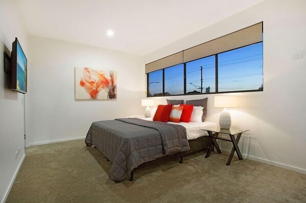 A bedroom with a bed, a nightstand, a lamp, a painting, and a large window.
