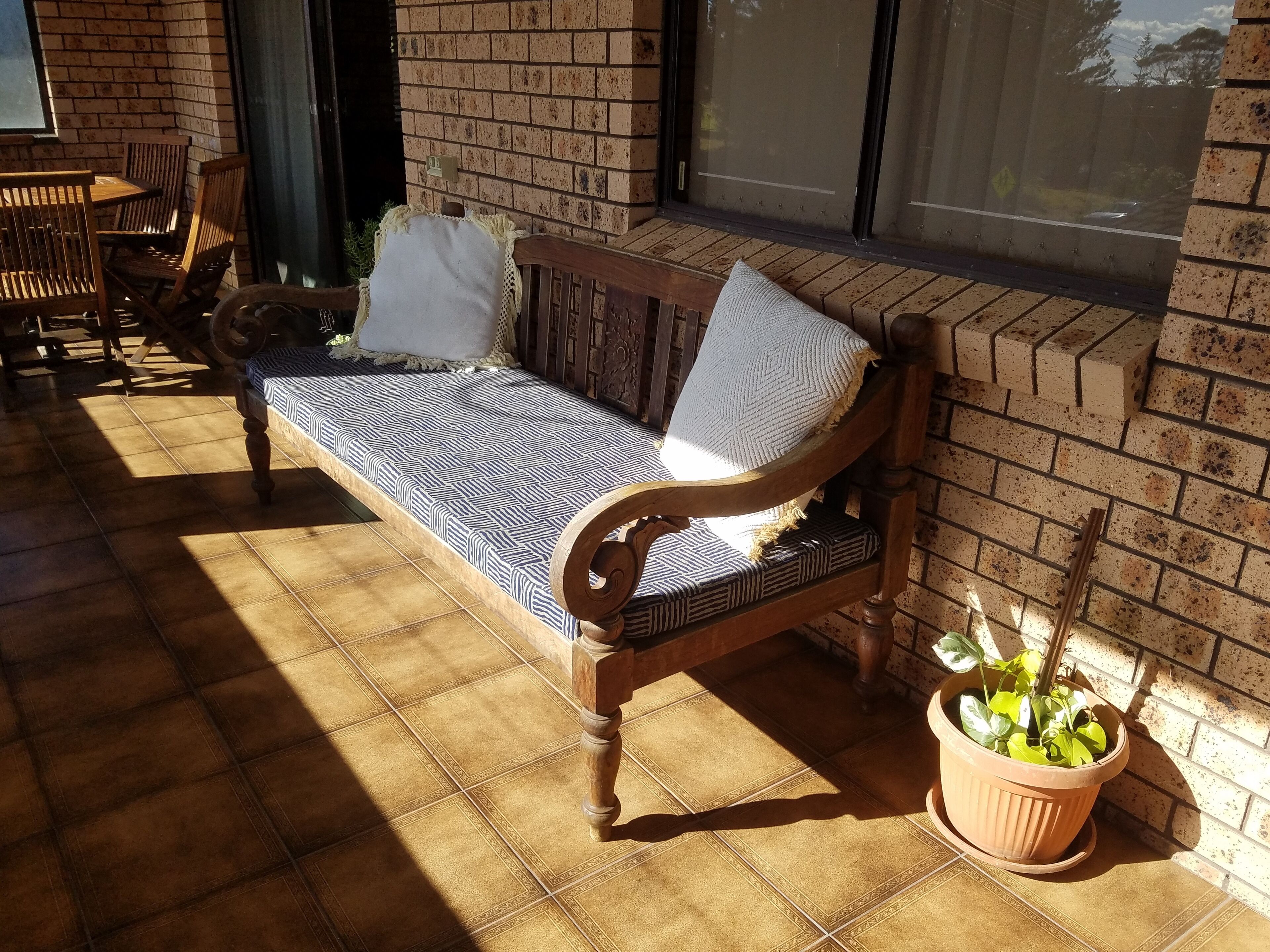 Un banco de madera con cojines estampados, una planta en maceta y un muro de ladrillo.