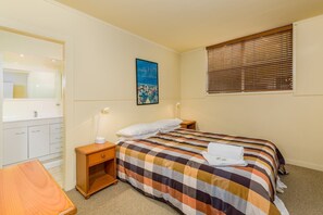 Ein Hotelzimmer mit kariertem Bettbezug, einer Nachttischlampe, einem Holznachttisch, einem kleinen Holztisch, einem Badezimmer mit Spiegel und Waschbecken sowie einem Fenster mit Jalousien.