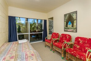 Une pièce comprenant un lit, deux fauteuils en osier avec un revêtement rouge, une grande fenêtre avec des rideaux et un tableau encadré accroché au mur.