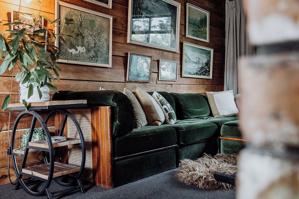 A green sofa with multiple cushions, a wooden table with a plant, and framed pictures on the wall.