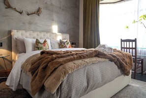 Une chambre à coucher avec une tête de lit capitonnée, un grand lit avec de nombreux oreillers, un plaid en fausse fourrure et une table en verre avec des chaises.