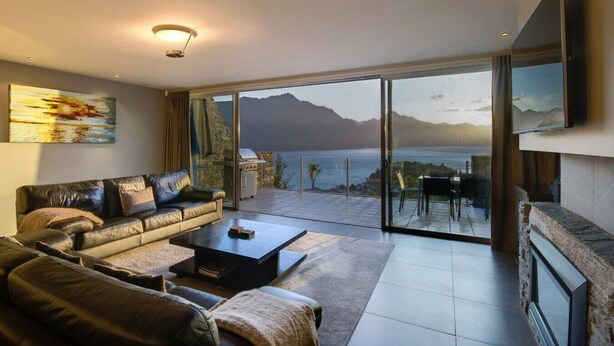 Un salon moderne avec un canapé en cuir, une table basse et une grande porte-fenêtre coulissante donnant sur un balcon avec vue sur la mer et les montagnes.