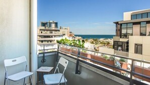 Un balcone con due sedie bianche e un tavolino, con vista su una zona costiera con edifici e un cielo azzurro intenso.