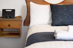Una cama bien hecha con un almohadón blanco y uno estampado, una mesita de noche con un dispositivo Sony y un cabecero de madera.