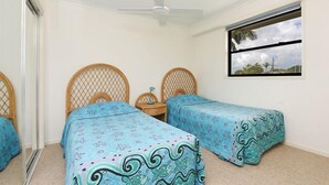 Un dormitorio con dos camas individuales, un ventilador de techo, una ventana con vista a palmeras y una mesita con una lámpara.