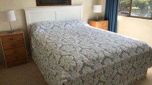 Un dormitorio con una colcha estampada, un cabecero blanco, una mesita de noche de madera con una lámpara y una ventana con cortinas.