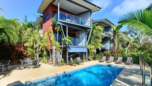Une maison moderne à deux étages, dotée d’un balcon, d’une piscine et d’un aménagement paysager de style tropical.