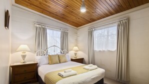 Ein ordentlich eingerichtetes Schlafzimmer mit Holzboden, zwei Nachttischen mit Lampen, einem großen Bett mit weißer und goldener Bettdecke und Vorhängen an den Fenstern.