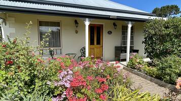 A two-story house with a corrugated metal roof, a front porch with a wooden door, and a garden with various plants and flowers.