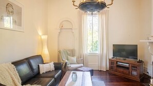 A living room with a black leather sofa, a wooden coffee table, a television on a wooden stand, a chandelier, and a window with curtains. - Bickley Valley Cottage (Bickley)