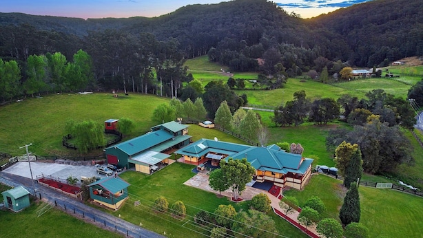 Vista aérea de una propiedad rural con varias construcciones, incluyendo una casa grande con techo verde, rodeada de campos y árboles verdes.
