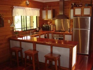 A kitchen with wooden cabinets, a stainless steel refrigerator, a stove, and a sink. There are three wooden stools around the bar area.