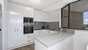 Una cocina moderna con armarios blancos, electrodomésticos de acero inoxidable y un amplio ventanal con vista a un edificio de ladrillo.