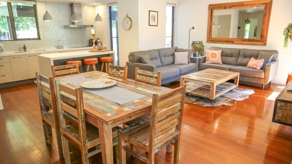 A modern living space with a wooden dining table, grey sofa, and a kitchen area with a sink and stools. - CHAPTER 11 DELANY: Bright (Bright)
